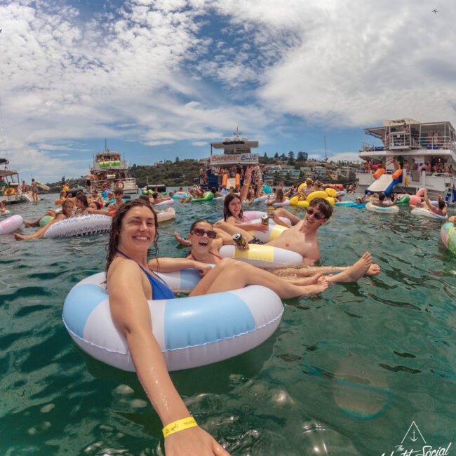 A group of smiling people relax in inflatable pool floats on the water during a lively outdoor event. Boats and more people on floats are visible in the background under a partly cloudy sky.