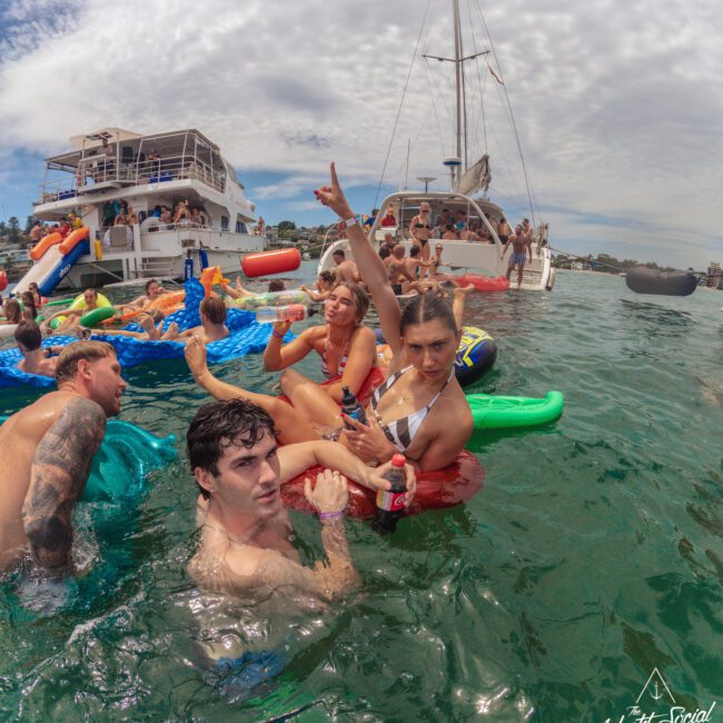 A group of young adults relax on colorful inflatables in the water near two yachts, holding drinks and enjoying a lively party under a partly cloudy sky. Some pose for the camera, surrounded by other partygoers.