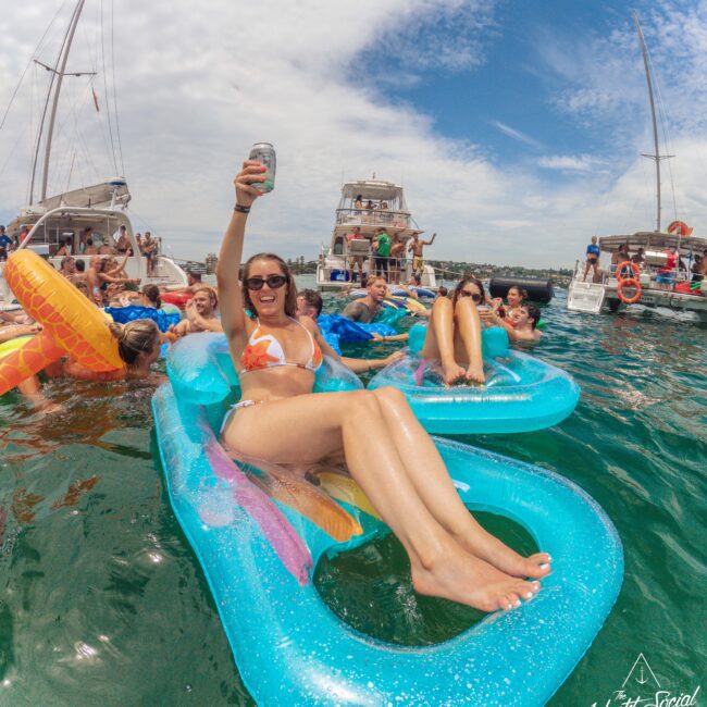 A group of people relax on colorful pool floats in the water near several yachts. A woman in sunglasses and a bikini raises a drink and smiles at the camera. The sky is partly cloudy and the scene is lively and festive.