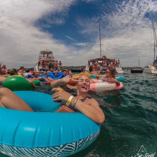 A young woman in a blue inflatable ring holds a drink and smiles at the camera while floating on the water. Other people in colorful floaties and boats are in the background under a partly cloudy sky.