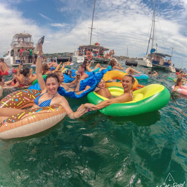 A group of people float on colorful inflatable pool toys in the sea, smiling and holding hands, with yachts and more people on inflatables in the background under a partly cloudy sky.