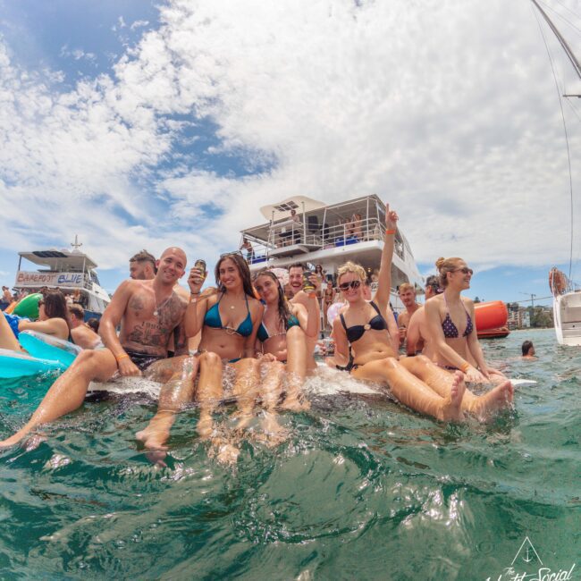 A group of people in swimwear sit on the edge of a boat, laughing and posing for a photo with the ocean and another boat in the background under a partly cloudy sky.