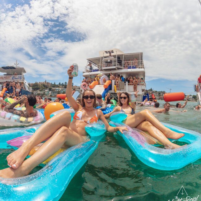 Two women in bikinis relax on blue pool floats in the water, smiling and holding drinks, surrounded by people and floats near a large yacht during a sunny pool party event.