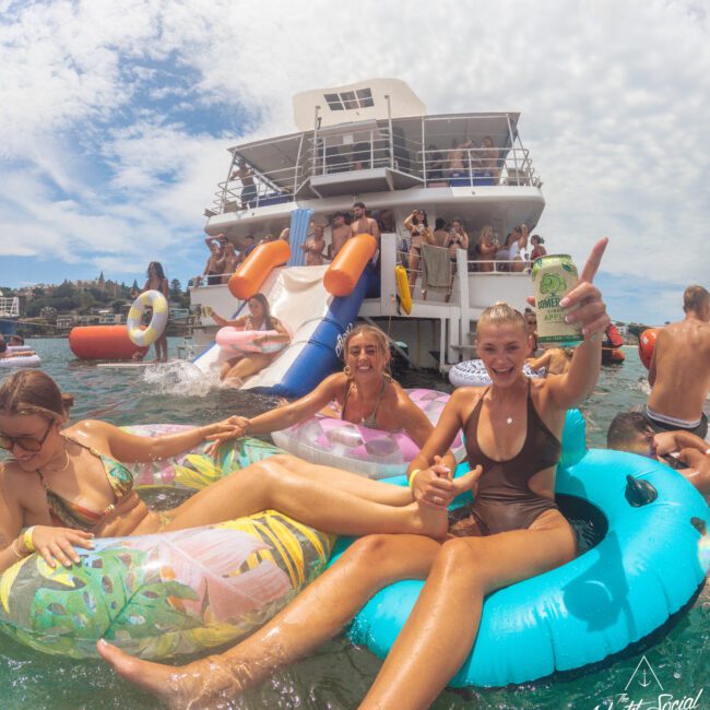 A group of young people relax on colorful pool floats in the water near a large white yacht, smiling and enjoying drinks. Others are partying and using a slide on the yacht under a bright, sunny sky.