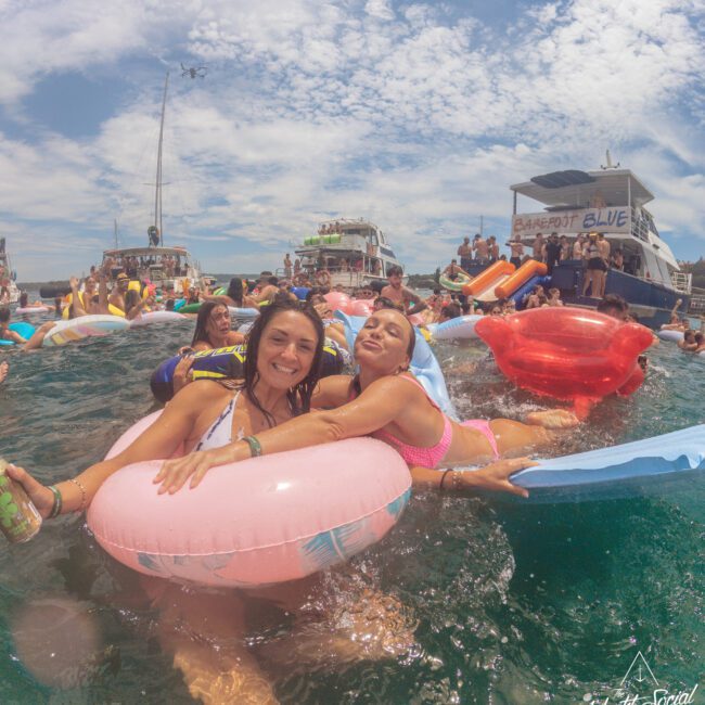 Two women smiling in pool floats on water, surrounded by people on various inflatables at a lively boat party; boats and blue sky are visible in the background.