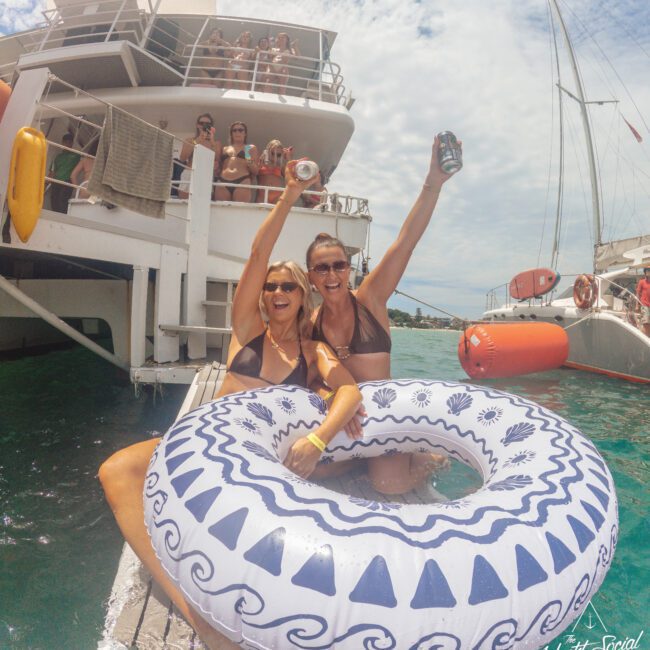 Two smiling women in swimsuits sit in a pool float, holding up drinks on a dock beside a boat. People on the boat cheer in the background under a sunny sky, enjoying a lively party on the water.