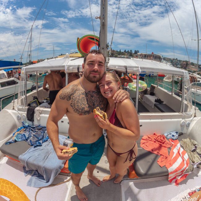 A smiling man and woman in swimsuits stand on a boat, holding sandwiches. Colorful towels and floaties are around them, and a sunny sky and water with other boats are visible in the background.