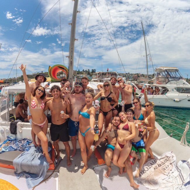 A group of friends in swimsuits smile, laugh, and pose for a photo on the deck of a boat on a sunny day, with water, boats, and a coastal town visible in the background.