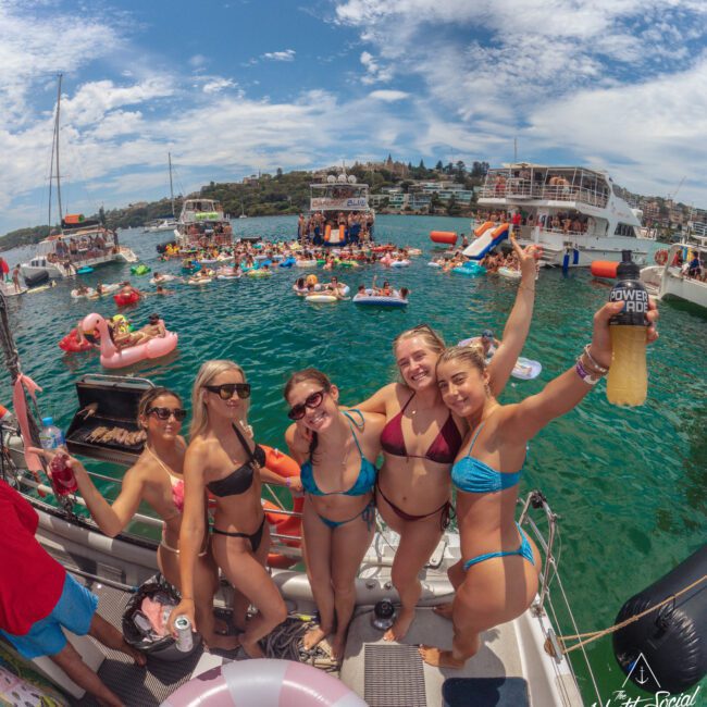 A group of five women in swimsuits smile and pose on a boat docked in a lively bay, with other people swimming, relaxing on inflatables, and boats nearby under a partly cloudy sky.