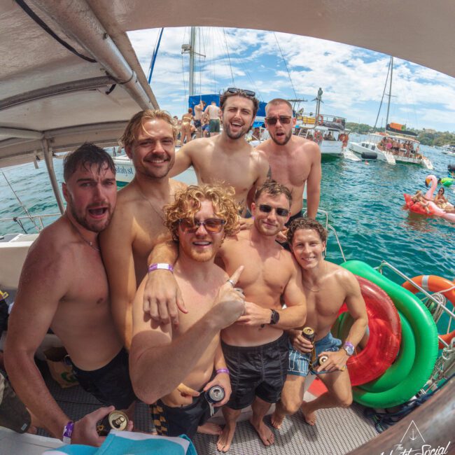 A group of seven smiling, shirtless men stand close together on a boat with drinks in hand, posing for a photo. Behind them are colorful pool floats and other boats on a sunny day at sea.