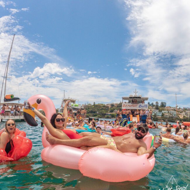 A group of people enjoy a lively party on the water, lounging on colorful inflatable floats, including a large pink flamingo. They smile, hold drinks, and relax under a sunny sky with boats and a city in the background.