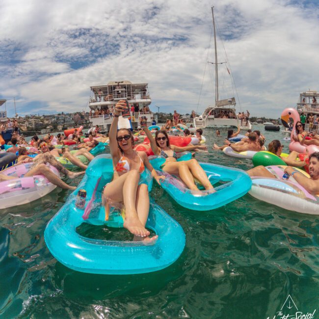 A group of people relax on colorful floaties in the water during a lively party with boats anchored nearby. Two women in sunglasses and swimsuits smile and toast the camera. The sky is partly cloudy.