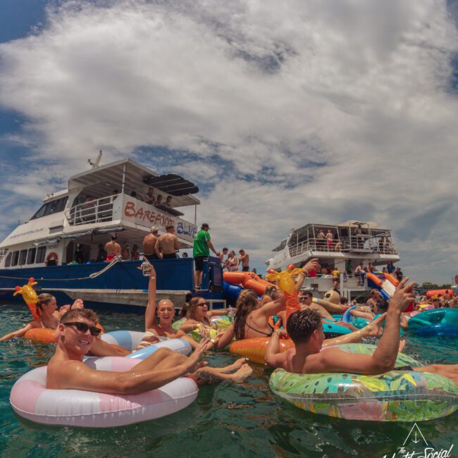 A group of people relax on colorful inflatable floats in the water near two large boats under a partly cloudy sky, enjoying a lively, social, summertime atmosphere.