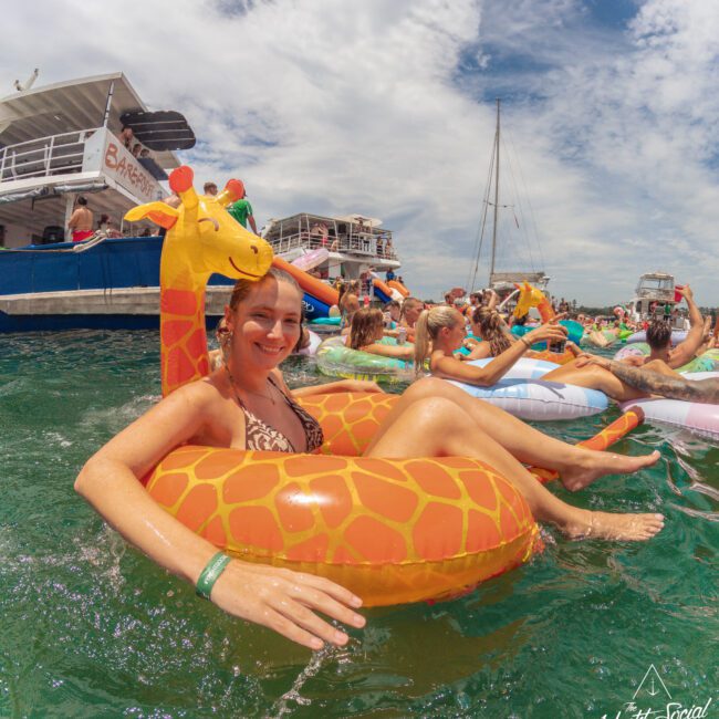 A smiling woman relaxes on a giraffe-shaped pool float in a crowded water party, surrounded by other people on inflatables. Boats and a partly cloudy sky are visible in the background.