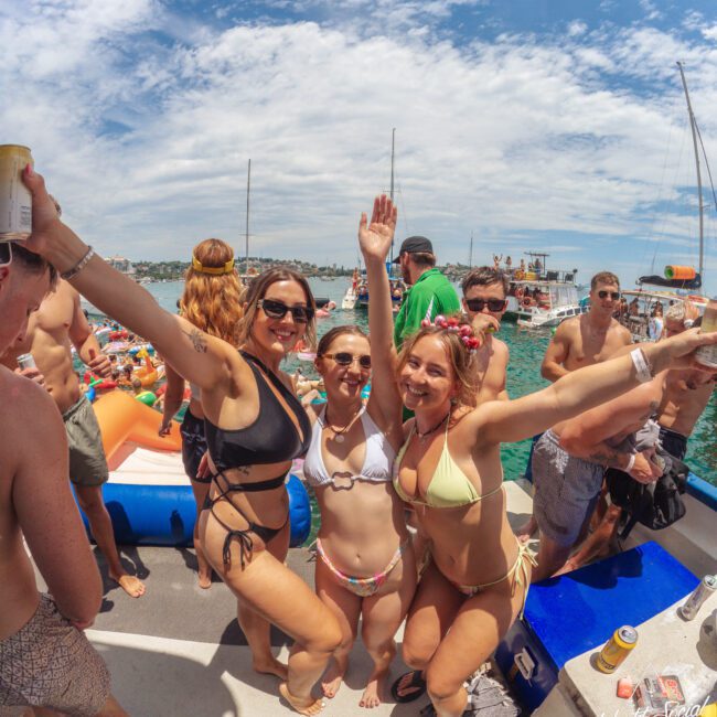 Three women in swimsuits pose and smile with arms raised on a crowded boat party, surrounded by other people, inflatables, and drinks, with boats and blue water in the background under a partly cloudy sky.