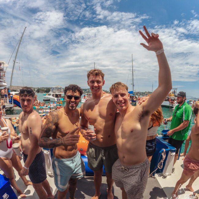 Four young men in swim trunks pose and smile at a lively boat party with many people, boats, and blue water in the background under a partly cloudy sky. One man raises his hand in celebration.