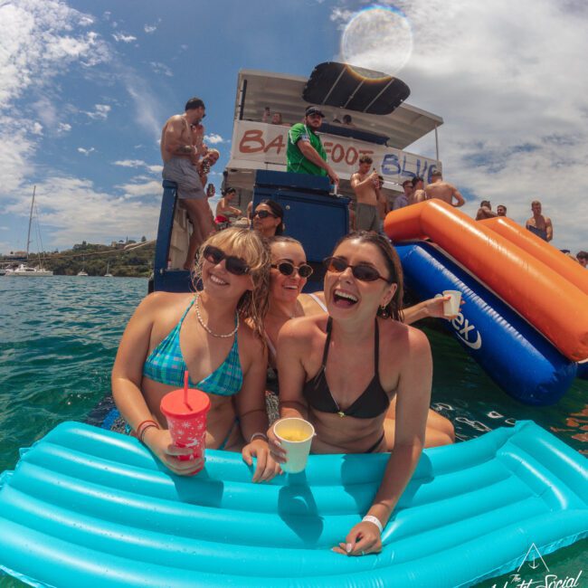 Three women in swimsuits smile and hold drinks while relaxing on a blue inflatable raft in the water. Behind them, people enjoy a party on a boat and an inflatable slide under a sunny sky.