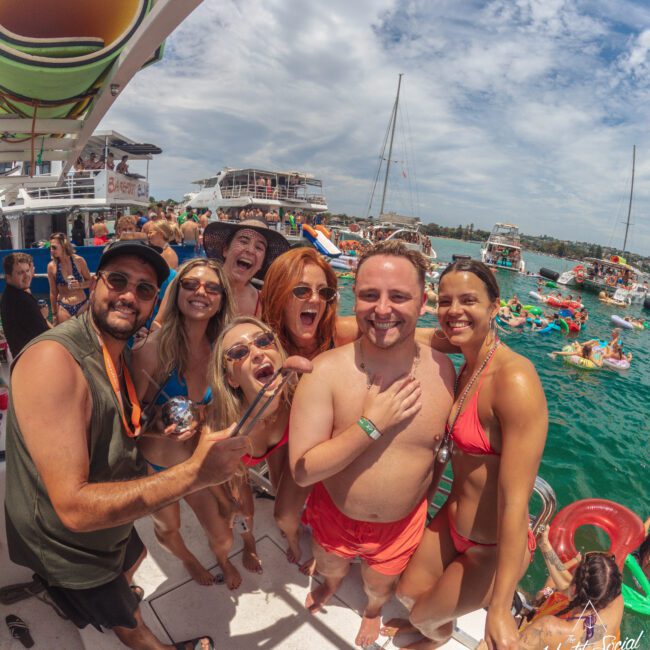A group of smiling people in swimsuits pose on a boat during a lively yacht party, with other boats, floating rafts, and partygoers in the water under a partly cloudy sky.