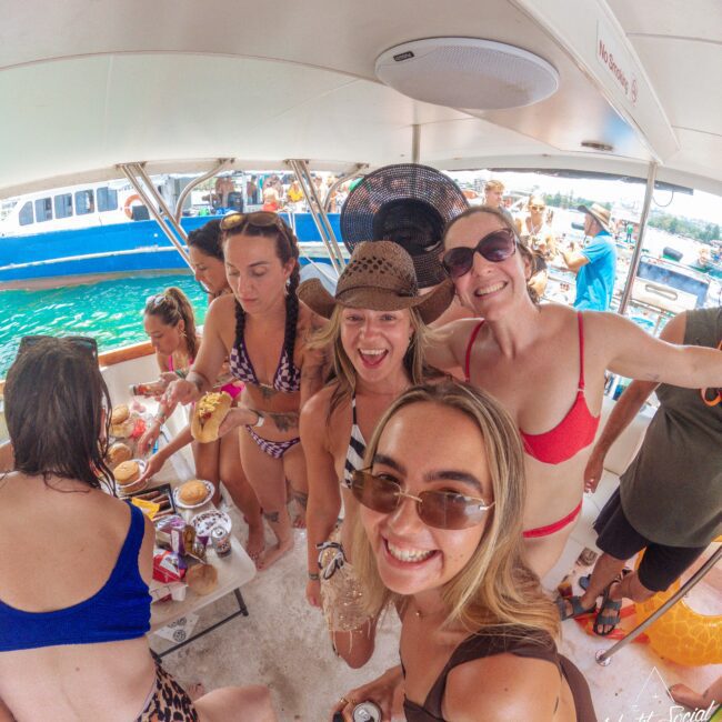 A group of women in swimsuits smile and pose for a selfie on a boat, surrounded by food and drinks, with turquoise water and another boat visible in the background.