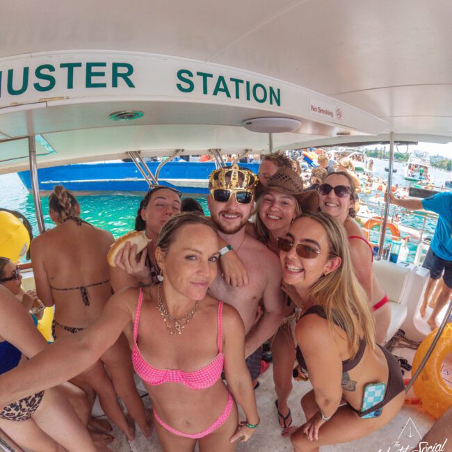 A group of smiling people in swimsuits gather for a selfie on a boat. One man wears a crown and sunglasses, surrounded by women. The boat is crowded and the water and other boats are visible in the background.