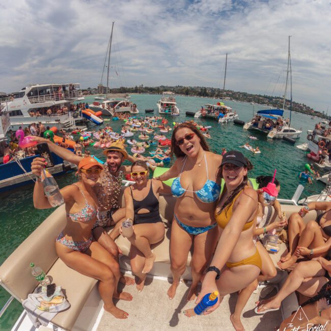 A group of people in swimsuits smile and pose on a boat surrounded by others partying in the water and on nearby boats, under a partly cloudy sky. The atmosphere is festive and summery.