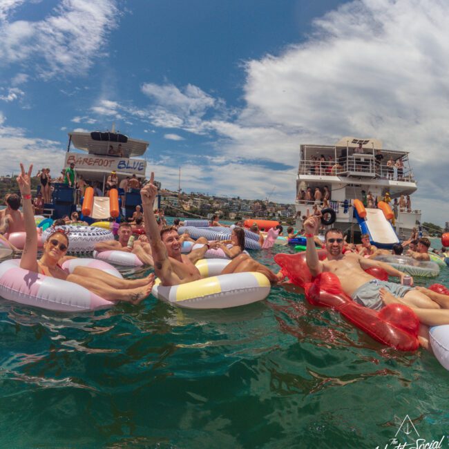 A group of people relax on colorful inflatables in the water near yachts, smiling and making peace signs, with a bright blue sky and city in the background. The atmosphere is festive and summery.
