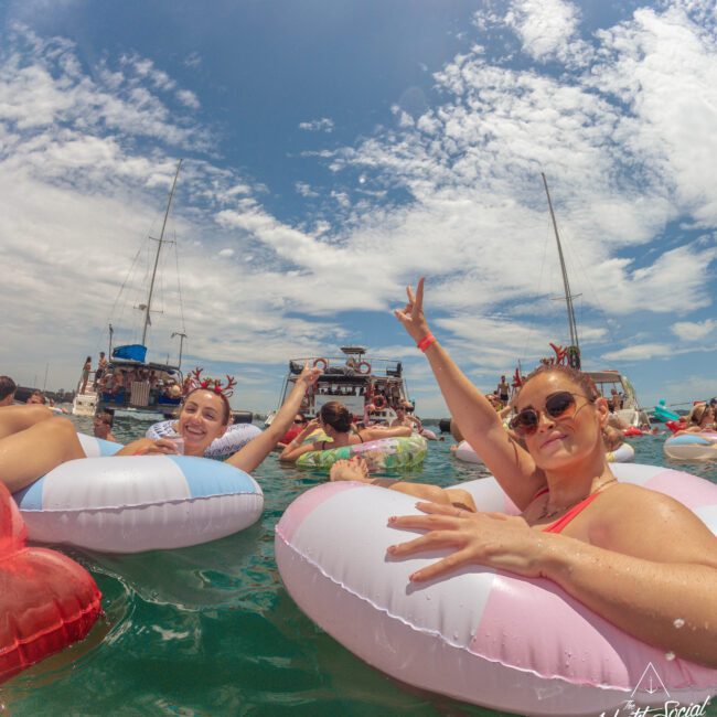 People relaxing on colorful pool floats in the ocean, wearing swimsuits and sunglasses. Boats float in the background under a partly cloudy sky. The atmosphere is festive and joyful, with some partygoers making peace signs.