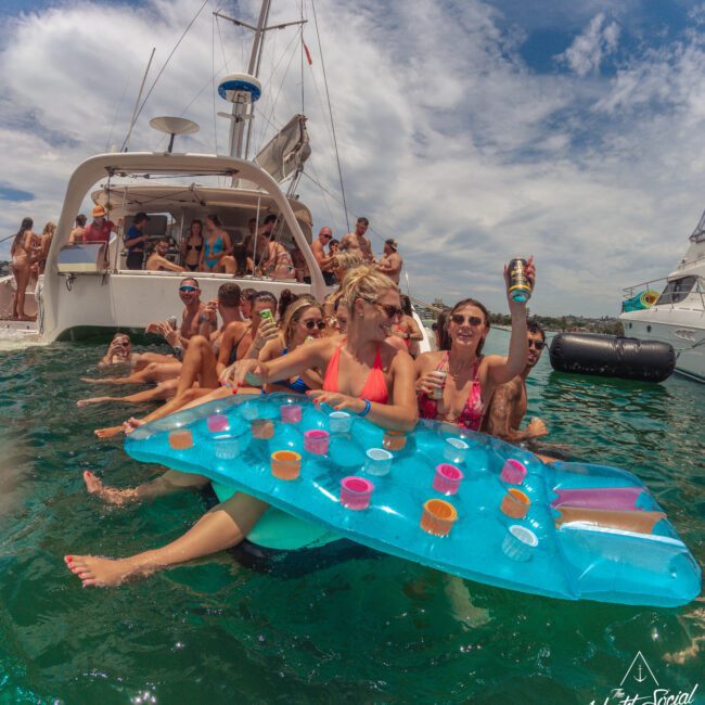 A group of people in swimsuits relax on a blue floating drink holder in the ocean near a yacht, smiling and raising drinks, with others enjoying the sun on the boat and in the water.