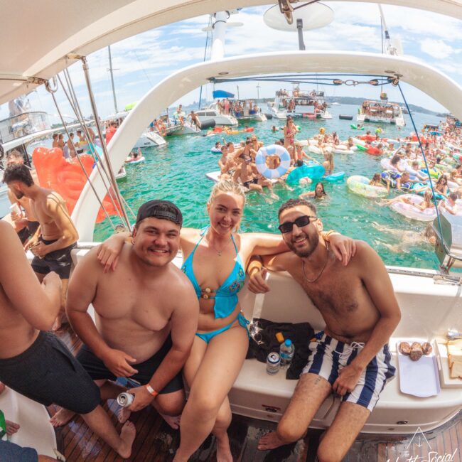 Three people in swimsuits smile and pose on a yacht during a lively pool party, with many others swimming and floating on colorful inflatables in the water around them on a sunny day.