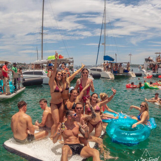 A group of young adults in swimwear smile and pose with drinks on a floating mat in the water, surrounded by yachts and other people enjoying a sunny party on the sea. The mood is festive and relaxed.