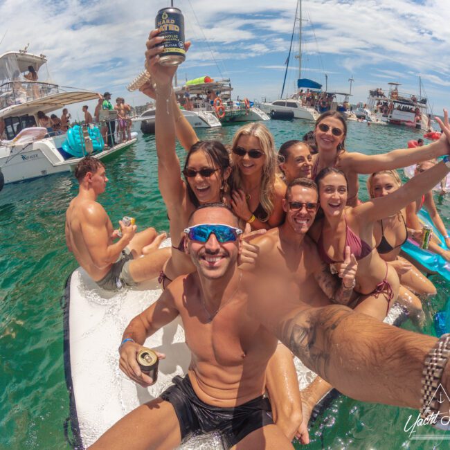 A group of friends in swimsuits smile and pose for a selfie on an inflatable raft in the water, surrounded by boats, during a sunny day at a lively yacht party.