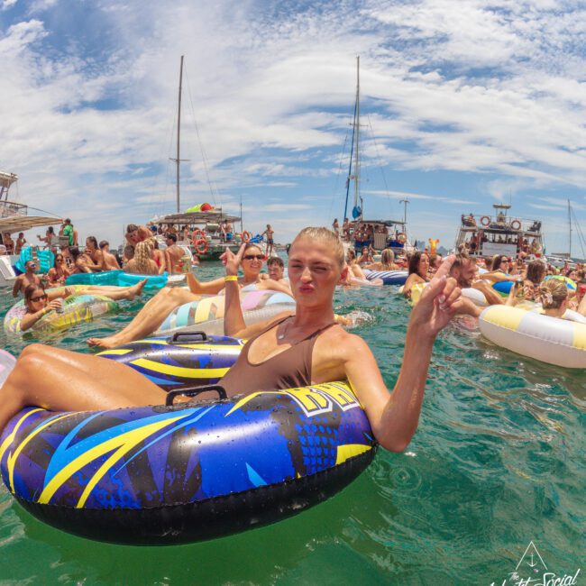 A woman in a swimsuit floats on an inflatable tube in the water, making a hand gesture and smiling at a lively crowd of people on floats, with boats and blue sky in the background.