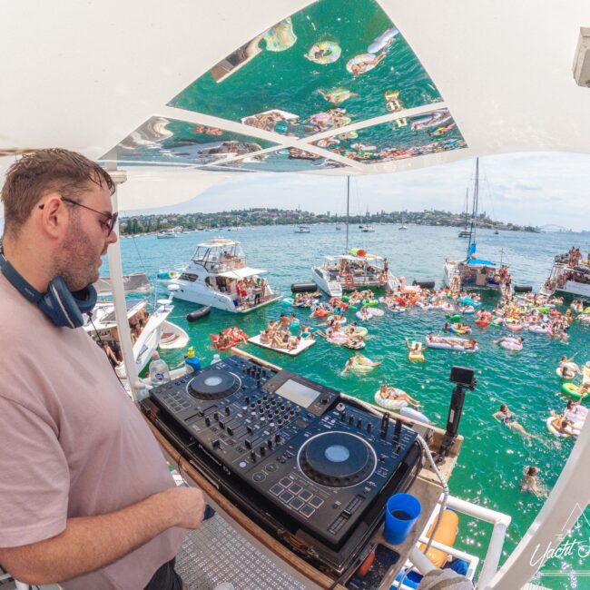 A DJ stands at a deck on a boat, playing music for a large crowd of people floating on inflatables in the water. Other boats and yachts are anchored nearby under a bright, sunny sky.
