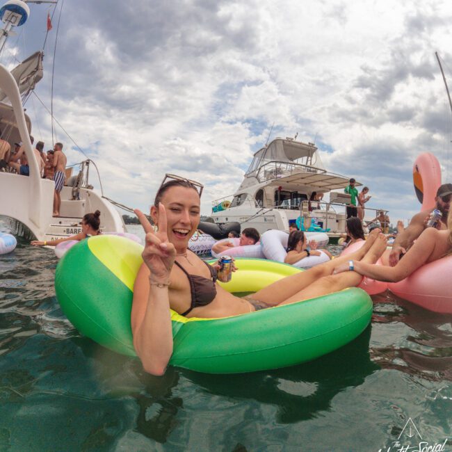 A smiling woman in a black bikini lounges on a green inflatable in the water, holding a drink and making a peace sign. Boats and other people on floaties are in the background under a cloudy sky.