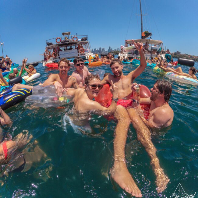 A group of young adults in swimsuits laughing and posing together on colorful pool floats in the ocean, surrounded by more people, boats, and a clear blue sky. The atmosphere is festive and lively.