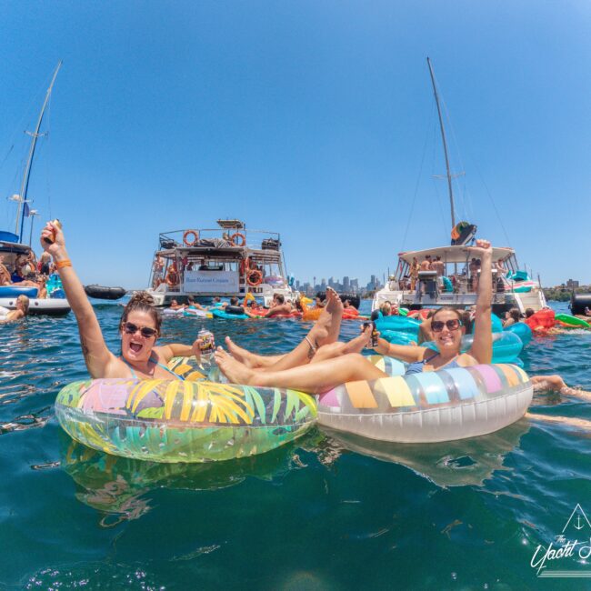 Two people float on colorful pool rings in the water, smiling and raising their arms, with boats and more people celebrating around them under a clear blue sky; city skyline visible in the background.
