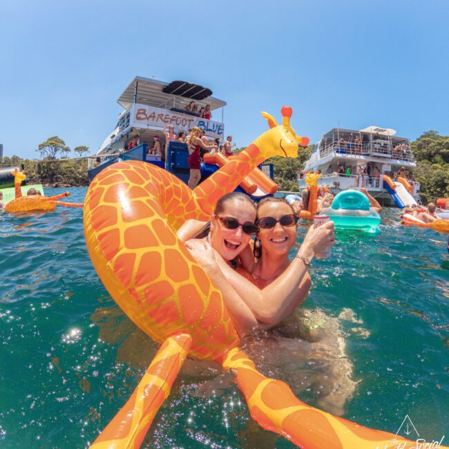 Two smiling women in sunglasses float on a large inflatable giraffe in clear blue water, with boats and other people enjoying the sunny day in the background.