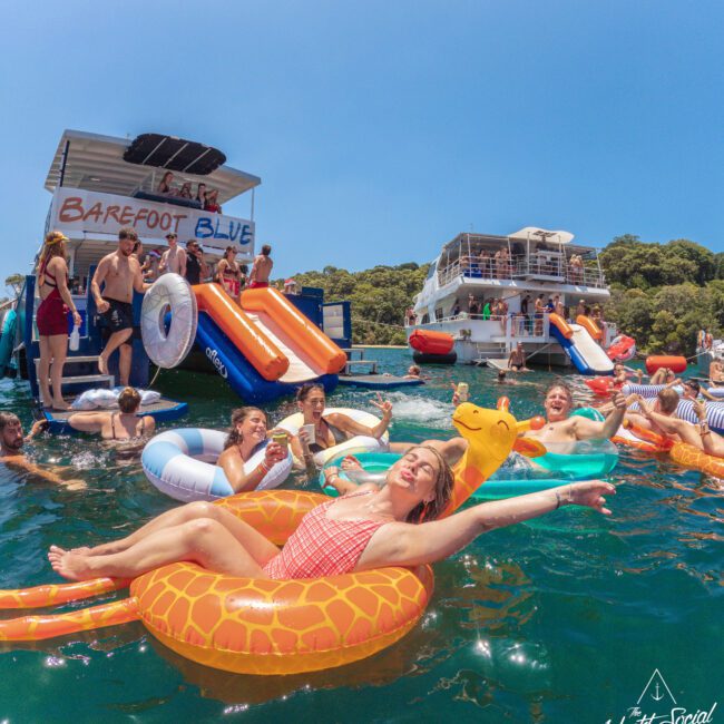 A group of people enjoy a lively yacht party, relaxing on colorful inflatable pool floats and slides in clear blue water near two large boats under a sunny sky. One person smiles and poses on a giraffe float in the foreground.