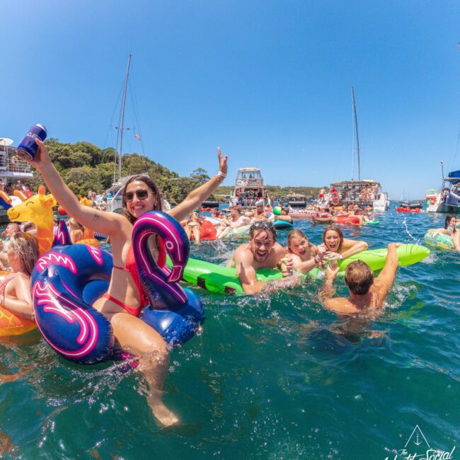 A group of people enjoy a sunny day on the water, lounging on colorful pool floats and swimming near anchored boats. The scene is lively and festive, with bright blue sky and clear green water.