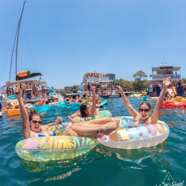 People relax on colorful pool floats in the water, raising their arms and smiling. Several boats with more partygoers are in the background under a clear blue sky. The scene is lively, festive, and summery.