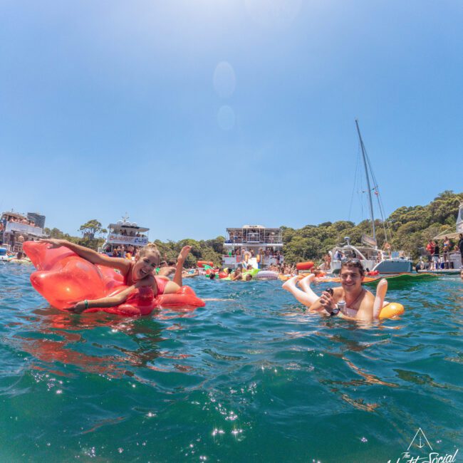 Two people float on inflatable pool toys in a busy, sunny harbor with boats and a crowd in the background. The water is blue, and the sky is clear. "Yacht Social Club" is written in the bottom right corner.