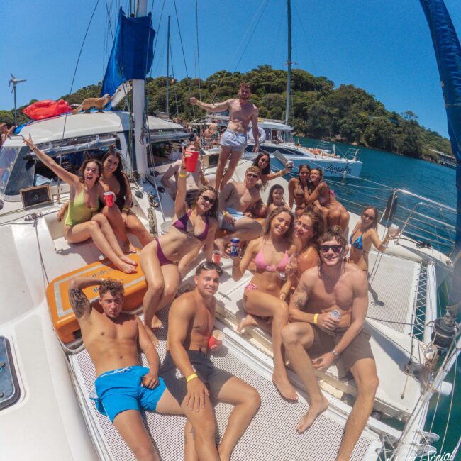A group of young adults in swimsuits relax and pose for a photo on the deck of a sailboat under a sunny sky, surrounded by blue water and lush green trees in the background.