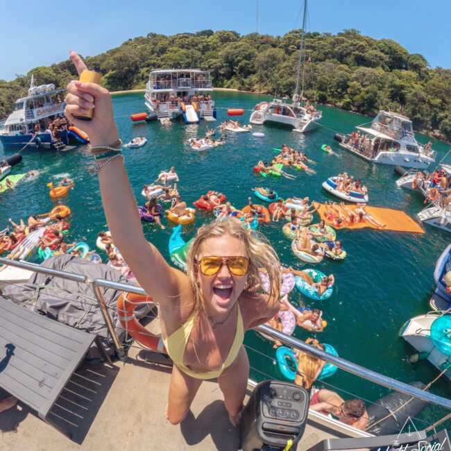 A smiling woman in a yellow bikini and sunglasses stands on a boat, giving a thumbs-up to the camera. Behind her, people lounge on colorful pool floats in the water near several boats anchored by a forested shore.