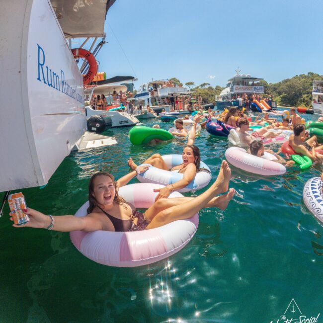 A group of people in colorful inflatable tubes relax and smile while floating on water near boats during a sunny yacht party. Some hold drinks and enjoy the lively, festive atmosphere.
