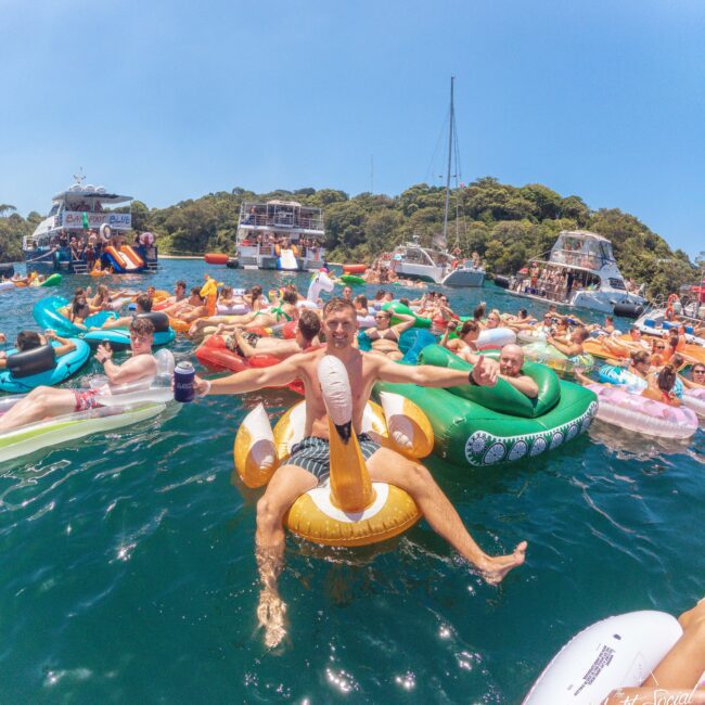 A group of people relax on colorful inflatable pool floats in the water, surrounded by boats, enjoying a sunny day. The scene is lively with many people, and green trees are visible in the background.