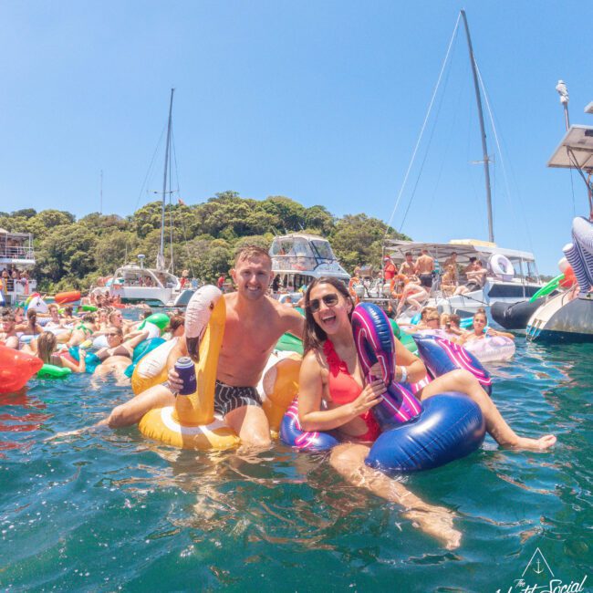 A man and woman laugh while sitting on colorful inflatables in the water, surrounded by people, boats, and yachts under a bright blue sky at a lively outdoor gathering.