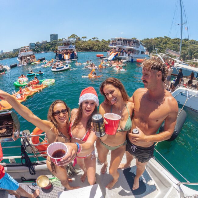 Four people in swimsuits on a boat, smiling and holding drinks. One wears a Santa hat. Other boats and people are visible in the water behind them, enjoying a sunny day.