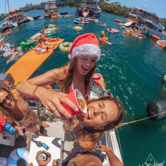 A woman in a Santa hat pours a drink into another woman’s mouth at a lively boat party. People in swimsuits relax on inflatables and nearby boats on a sunny day, with city buildings and trees in the background.