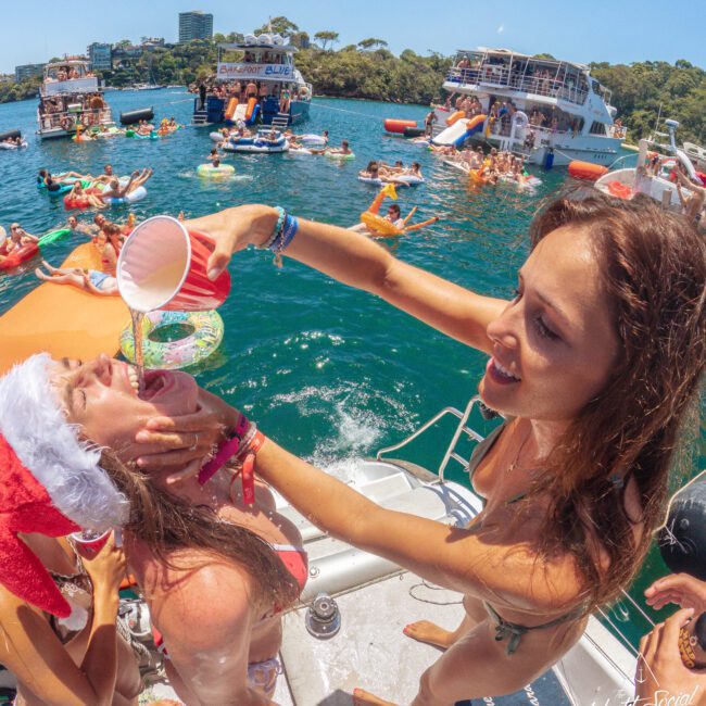 A woman in a bikini pours a drink into another woman's mouth, who is wearing a Santa hat, at a lively boat party on the water with many people, floaties, and boats in the background.