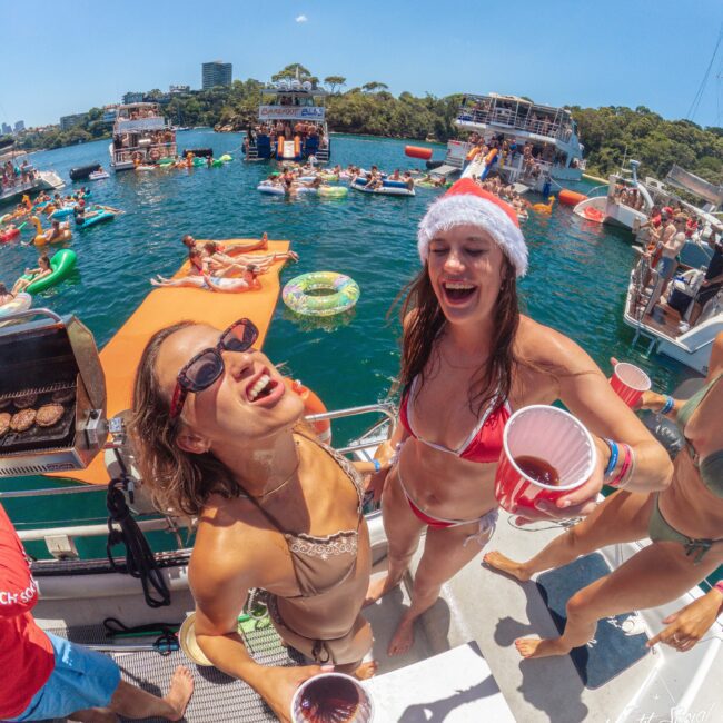 Two women in swimsuits, one wearing a Santa hat, smile and hold red cups on a boat during a lively summer party. Other people are swimming, lounging on floats, and relaxing on nearby boats in sunny weather.